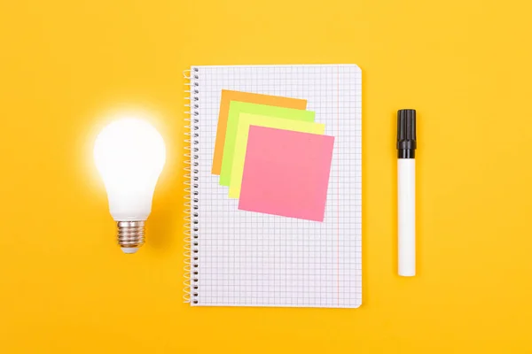 Glowing Power Saving Lamp and Notepad with Sticky Notes Lying on Yellow Table, Blank Template with Copy Space - Top View, Flat Lay