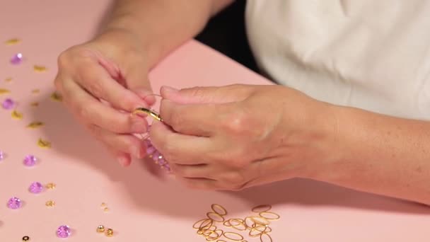 手工珠宝 身穿白色T恤的欧洲白人妇女在家里的粉红桌上手工制作Bijouterie Beads和Bracelets 工匠工作场所 特写镜头 静态镜头 — 图库视频影像