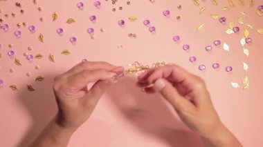 El yapımı Mücevher. Beyaz Tişörtlü Beyaz Avrupalı Kadın Pembe Masa 'da El yapımı Bijouterie, Boncuk ve Bilezik Yapıyor. Sanatçılar İş Yeri - Üst Görünüm, Sabit Çekim