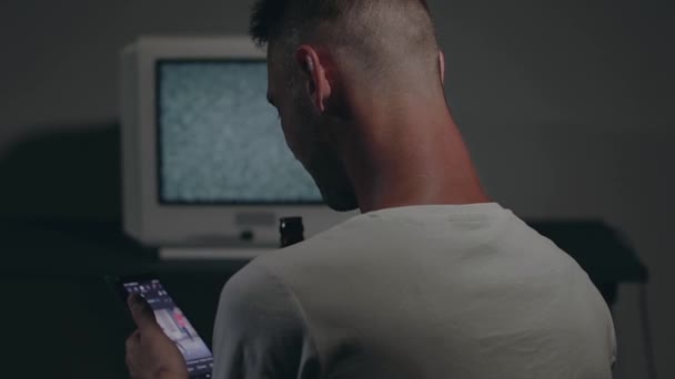 白いTシャツの男が暗室の古いシルバーテレビに向かって座っている ビールを飲み スマートフォンを使う テレビ信号を検索しましょう 画面上の歪み バックビュー 中程度のクローズアップ フィルムグレイン — ストック動画