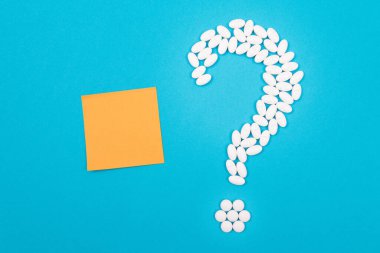 Beyaz haplardan ve tabletlerden yapılan soru işareti küçük boş not kağıdıyla, mavi arka planda yatıyor. Global İlaç Endüstrisi ve Tıbbi Ürünler