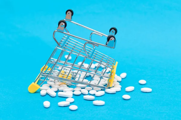 Buying Medicines. Expensive Medicine and Inflation: Scattered Pills or Capsules and Shopping Cart on Blue Background. Global Pharmaceutical Industry and Big Pharma. Ordering Pharmaceutical Products