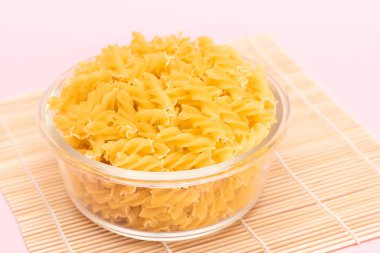 Cam kavanozda pişmemiş Fusilli Makarna, pembe arka planda Bambo Mat üzerinde. Şişman ve Sağlıksız Yemek. Klasik kuru sarmal makarna. İtalyan Kültür ve Mutfağı. Çiğ Makarna
