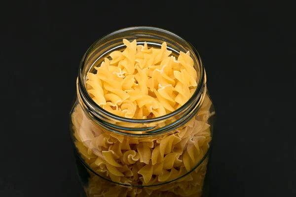 Uncooked Fusilli Pasta in Glass Jar on Black Background. Raw and Dry Macaroni. Unhealthy and Fat Food. Italian Culture