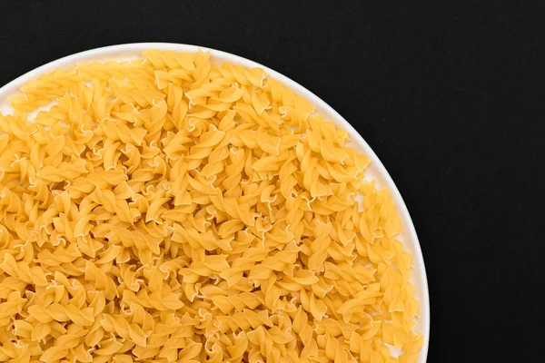 Pişmemiş Fusilli Makarna Siyah Arkaplanda Beyaz Tabakta Yatıyor. Çiğ ve kuru makarna. Sağlıksız ve Yağlı Gıda - Üst Görünüm