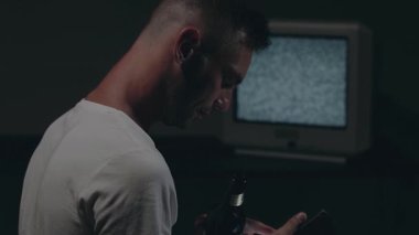 Beyaz tişörtlü adam Karanlık Oda 'da Eski Gümüş TV' ye Karşı Oturuyor. Bira içip, akıllı telefonunu kullanmaktan başka. Televizyon sinyalini ara. Ekrandaki bozulma. Arka Görünüm, Orta Yakın Çekim, Film Tanesi