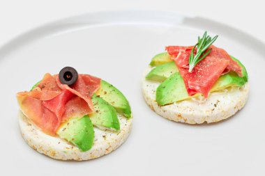 Beyaz Tabakta Avocado, Jamon, Zeytin ve Rosemary ile Rice Cake Sandviçi. Kolay Kahvaltı 'da. Hızlı ve sağlıklı sandviçler. Lezzetli dolgulu kızarmış ekmek. Sağlıklı Beslenme Atıştırması - İzolasyon