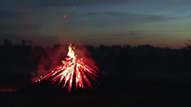 Big Burning Campfire in Early Morning or Evening against the Blue Sky. Wood on Fire. Flying Sparks. Travel and Tourism Concept. Giant Flaming Bonfire at Summer - Static Shot, Slow Motion