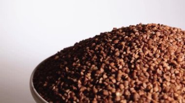 Dry Uncooked Brown Buckwheat Groats Heap on White Plate Rotating against White Background. Raw Large Buckwheat Grains. Russian, Ukrainian and Belarusian Culture