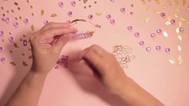 El yapımı Mücevher. Beyaz Tişörtlü Beyaz Avrupalı Kadın Pembe Masa 'da El yapımı Bijouterie, Boncuk ve Bilezik Yapıyor. Sanatçılar İş Yeri - Üst Görünüm, Sabit Çekim