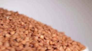 Dry Uncooked Brown Buckwheat Groats Heap on White Plate Rotating against White Background. Raw Large Buckwheat Grains. Russian, Ukrainian and Belarusian Culture