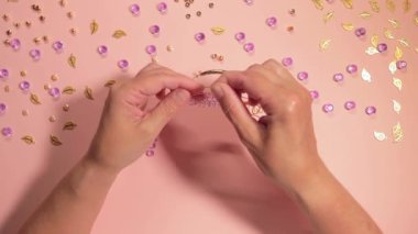 El yapımı Mücevher. Beyaz Tişörtlü Beyaz Avrupalı Kadın Pembe Masa 'da El yapımı Bijouterie, Boncuk ve Bilezik Yapıyor. Sanatçılar İş Yeri - Üst Görünüm, Sabit Çekim