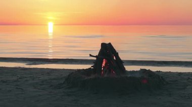 Sakin Deniz 'e karşı şenlik ateşi ya da Okyanus ve Akşam Güneşinin batışı - Statik Çekim, Yavaş Hareket. Kamp ateşi Kum üzerinde yanıyor. Ateşe odun. Şenlik Ateşi için yer. Deniz kenarındaki ateş çukuru. İşaret için Ateş