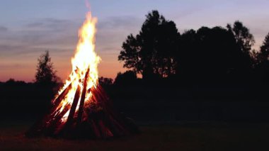 Big Burning Campfire at Summer Evening against the Blue Sky. Wood on Fire. Flying Sparks. Travel and Tourism Concept. Giant Flaming Bonfire - Static Shot, Slow Motion
