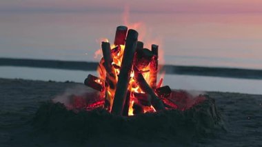 Plajda Sakin Deniz 'e karşı şenlik ateşi ya da Karanlık Romantik Akşam' da Okyanus - Sabit Çekim, Yavaş Hareket. Kamp ateşi Kum üzerinde yanıyor. Ateşe odun. Şenlik Ateşi için yer. Deniz kenarındaki ateş çukuru. İşaret için Ateş