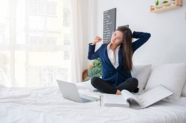 Overworked and Stressed young Asian woman working on laptop computer and document paper in bed at hotel, Business trip, Burnout, technology, copy space, Professional Burnout And Exhaustion Concept.