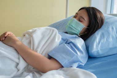 Üzgün Asyalı hasta kadın hastane yatağında yatıyor ve koronavirüsü korumak için maske takıyor. Sağlık hizmeti kavramı, karantina koronavirüsü (COVID-19) ve sağlık sigortası.