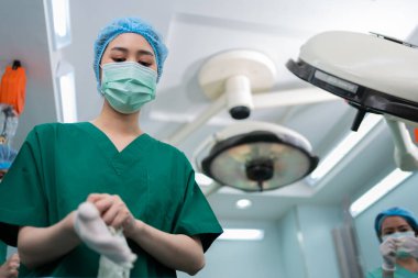 Tıbbi maskeli Asyalı cerrahın portresi ayakta duruyor ve hastane ameliyathanesinde tıbbi eldiven giyiyor. Profesyonel cerrahlar takımı. Sağlık hizmeti, acil tıbbi servis kavramı