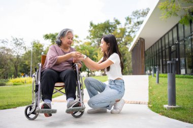 Asyalı dikkatli bakıcı ya da hemşire hastanın elini tutup hastayı tekerlekli sandalyeye oturtmaya teşvik ediyor. Bakıcının, birikimin ve sağlık sigortasının özenle mutlu emeklilik kavramı.