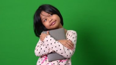 7-8 yıllık şirin Asyalı kız öğrenci portresi kameraya bakıyor ve yeşil ekran arkaplanı üzerine kitap tutuyor ve yeni fikirler düşünüyor. Akıllı çocuk kağıt kitaplarla öğrenmeyi seviyor..