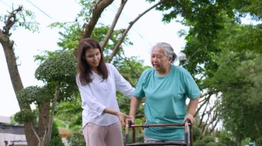 Yaşlı bir Asyalı kadın yürüteç kullanıyor ve kızıyla bahçede yürüyor. Mutlu emeklilik kavramı. Bakıcının, birikimin, sağlık sigortasının ve sağlık hizmetlerinin bakımıyla.