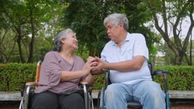 Mutlu Asyalı yaşlı çift tekerlekli sandalyede oturuyor ve yaşlı kadın koca elini tutuyor ve birlikte konuşuyor ve parkta dinleniyor emeklilerin boş vakitleri, yaşlı sağlık hizmetleri konsepti..