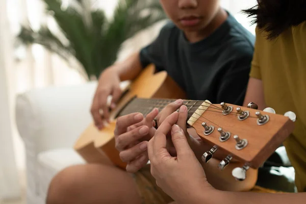 Asian boy playing guitar with mom in the living room for teaching him son play guitar, feel appreciated and encouraged. Concept of a happy family, learning and fun lifestyle, love family ties
