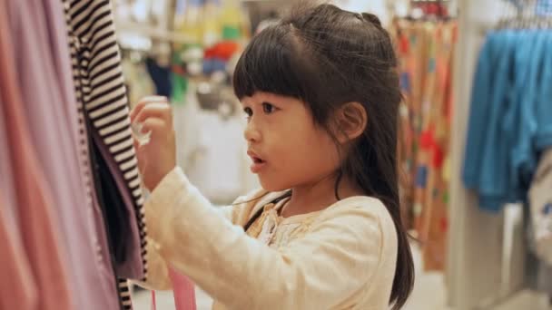 在一家购物中心的儿童时装店里 一个可爱的小女孩在许多衣服中挑选一件新衣服 销售概念 假日美及时尚概念 — 图库视频影像