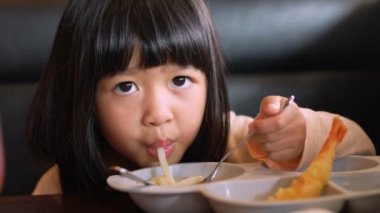 Restoranda oturan Asyalı kız ailesiyle birlikte Japon eriştesi (Udon veya ramen) ve Karides Tempura yiyor ve yemekten sonra baş parmağını gösteriyor. Lezzetli çok lezzetlidir. Çocukluk, sağlık konsepti.