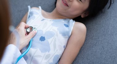 Asyalı kadın pediyatrist doktor muayene için stetoskop tutuyor küçük bir kız hasta ve çocuğun kalp ciğerleri, iyi aile doktoru çocuğu evde ziyaret ediyor, sağlık ve çocukluk konsepti için ilaç alıyor..