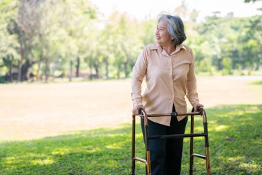 Yaşlı mutlu Asyalı kadın parkta osteoartrit rehabilitasyon fizyoterapisi için bir aylak kullanıyor. Bakıcının, birikimin ve sağlık sigortasının özenle mutlu emeklilik kavramı