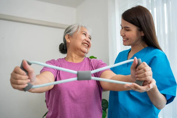 在物理治疗中心 老年女性快乐地与年轻理疗师一起使用橡胶膨胀机进行运动 以恢复手臂肌肉 在照顾者和健康的照料下快乐退休的概念 免版税图库图片