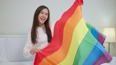 Pencere kenarındaki gökkuşağı bayraklarıyla yataklarda oturan mutlu Asyalı lezbiyenlerin portreleri. LGBTQ Yaşam Tarzı Gurur Ayı