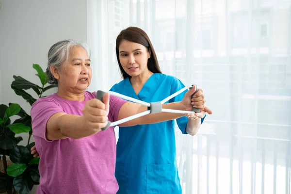 在物理治疗中心 老年女性快乐地与年轻理疗师一起使用橡胶膨胀机进行运动 以恢复手臂肌肉 在照顾者和健康的照料下快乐退休的概念 图库图片