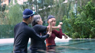 Young trainer helping senior woman in aqua aerobics and working out in the pool. old woman and mature man doing aqua aerobics exercise in swimming pool, Elderly sports, and active lifestyle concept.