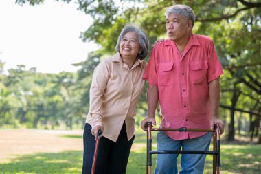 Yaşlı bir Asyalı adam yürüteç kullanıyor ve karısıyla parkta yürüyor. Aile ve bakıcı, birikim ve sağlık sigortası ile mutlu emeklilik kavramı