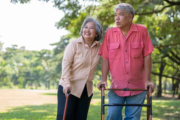 Yaşlı bir Asyalı adam yürüteç kullanıyor ve karısıyla parkta yürüyor. Aile ve bakıcı, birikim ve sağlık sigortası ile mutlu emeklilik kavramı