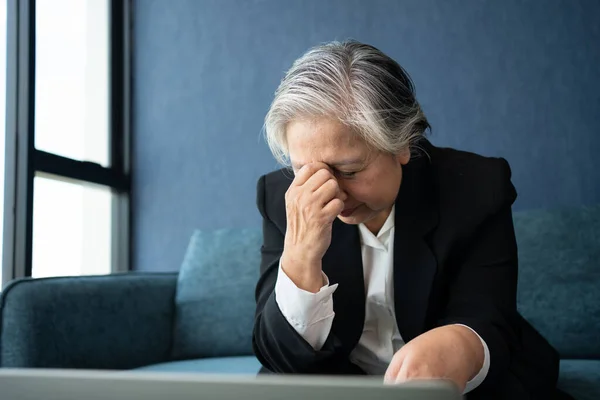 Overworked tired old lady holding head feeling headache. Asian senior business woman stress from hard work. Frustrated business woman thinks seated at workplace, feel distressed looks helpless concept