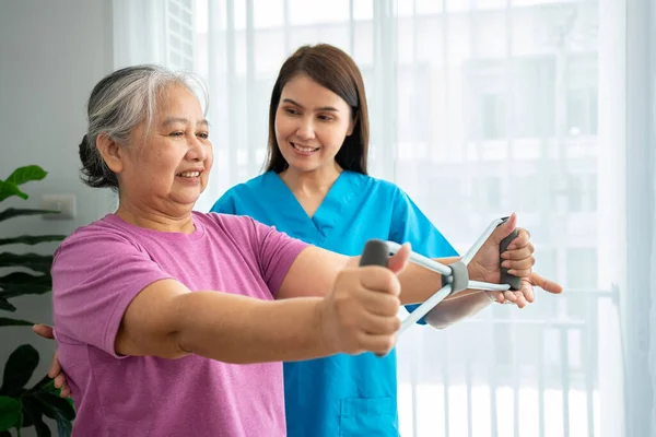 在物理治疗中心 老年女性快乐地与年轻理疗师一起使用橡胶膨胀机进行运动 以恢复手臂肌肉 在照顾者和健康的照料下快乐退休的概念 免版税图库照片