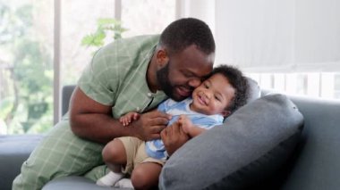 Mutlu Afrikalı Amerikalı adam oturma odasındaki kanepede küçük bebek bebekle oynuyor. Ebeveyn ve küçük çocuk evde dinleniyor. Ebeveynler ve küçük kız çocukları evde vakit geçirmekten zevk alıyorlar. Çocuk Bakımı