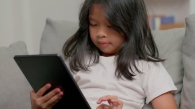 Küçük kız oturma odasında kanepede uzanmış elinde tabletle ekrana bakıyor ve çizgi film seyrediyor. Sadece dijital cihazda online uygulama kullanan bir çocuk. Çocuk ve elektronik cihazlar ve siber dolandırıcılık kavramı.