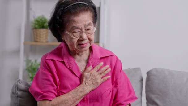 アジアの高齢のおばあさんは 心臓の痛みに苦しみ リビングルームのソファーに座っている間 胸を抱えていました 医療緊急治療と医療保険の保護コンセプト — ストック動画