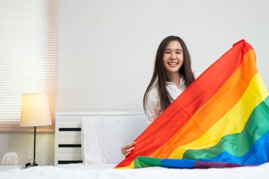 Pencerenin yanındaki yatağın üzerinde gökkuşağı bayraklarıyla oturan mutlu Asyalı lezbiyen kızların portreleri. İlişki konseptiyle yaşayan neşeli genç kadınlar. LGBTQ Yaşam Tarzı Gurur Ayı