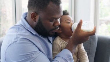Afrikalı baba kanepede oturuyor, kucağında yeni doğmuş bebeği oturma odasında süt şişesiyle besliyor. Otantik yaşam tarzı gerçek bir an. Bekar baba aile hayatı konsepti..