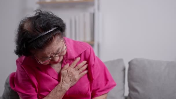 アジアの高齢のおばあさんは 心臓の痛みに苦しみ リビングルームのソファーに座っている間 胸を抱えていました 医療緊急治療と医療保険の保護コンセプト — ストック動画