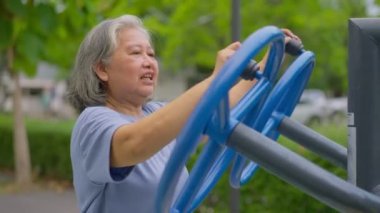 Kıdemli Asyalı kadın halka açık parktaki egzersiz istasyonlarında günlük antrenman yapıyor. Fiziksel aktivitelerle genç kalmayı, açık hava spor salonunu temsil ediyor. Fiziksel sağlık konsepti. Şehir parkında açık hava fitness ekipmanları