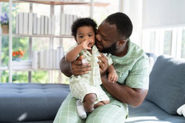 Mutlu Afrikalı Amerikalı adam oturma odasındaki kanepede küçük bebek bebekle oynuyor. Ebeveyn ve küçük çocuk evde dinleniyor. Ebeveynler ve küçük kız çocukları evde vakit geçirmekten zevk alıyorlar. Çocuk Bakımı