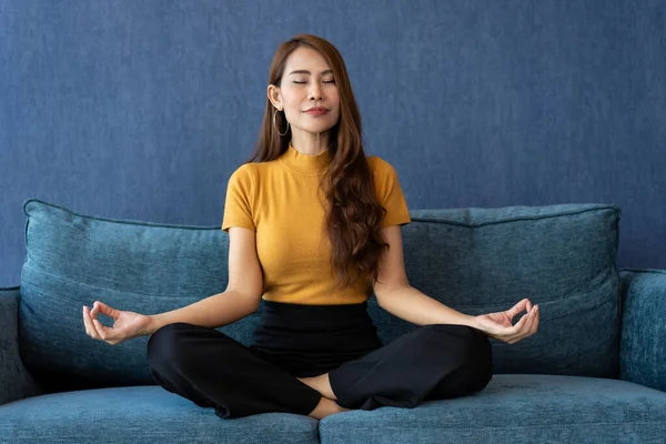 Portrait of healthy Asian woman meditating and exercise pose yoga on the sofa in morning. Zen relaxation and mindfulness lifestyle, Concept of indoor workout for good healthy and breathing practice.