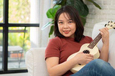Mutlu Asyalı kadın oturma odasında kanepede oturuyor ve rahatça ukulele çalışıyor. Sanatçıdan rahatlama ve ilham. Müzik kavramı, rahatlama ve hobi aktiviteleri boş zamanların tadını çıkar