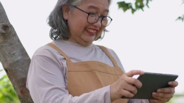 İnternet tableti kullanan mutlu Asyalı son sınıf öğrencisi kadın ulusal parkta dinlenirken yolculuğunu nasıl yapacağını araştırıyor. Rahat bir emeklilik yaşam tarzı ve iletişim kavramı.